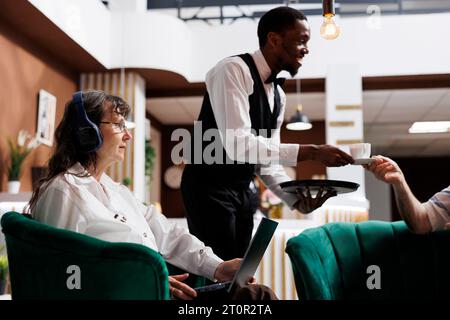 Tout en se relaxant dans le salon avec café et électronique, le personnel aimable de l'hôtel aide les couples âgés à s'enregistrer. Alors que la vieille femme à la retraite utilise un ordinateur portable, un employé afro-américain sert un homme âgé. Banque D'Images