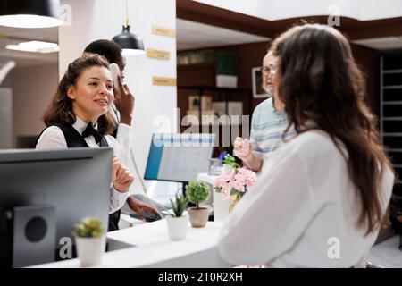 Touriste féminine âgée au comptoir du salon faire la réservation de la chambre pour les vacances. Une femme de tourisme retraitée s'enregistre à la réception avec l'aide de la réceptionniste, avide de vacances agréables. Banque D'Images