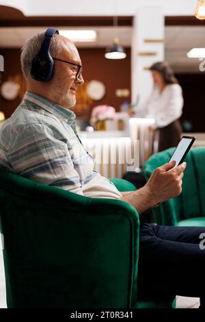 Homme caucasien âgé porte des écouteurs et est assis sur le canapé du hall de l'hôtel saisissant la tablette. Invité masculin senior ayant casque sans fil attend dans le salon et utilise gadget numérique pour naviguer sur Internet. Banque D'Images