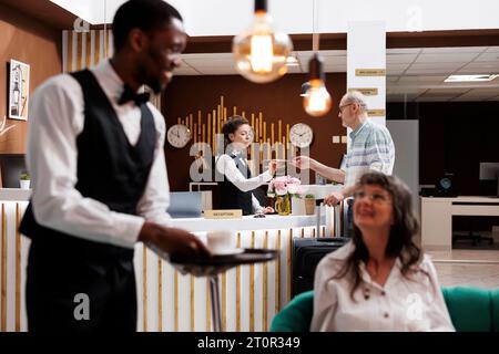 Arrivée à la réception de l'hôtel, client âgé s'enregistre avec l'assistance de la réceptionniste à la réception. La conciergerie féminine caucasienne reçoit un document personnel du vieil homme pour une inscription en douceur. Banque D'Images