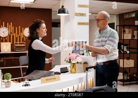 Une personne âgée de race blanche arrive à la réception de l'hôtel et s'enregistre avec un passeport. Au comptoir d'enregistrement, concierge féminin recevant un document d'identité du voyageur masculin senior pour la réservation de la chambre. Banque D'Images