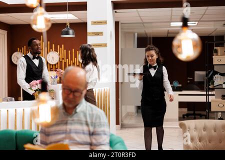 Femme âgée retraitée assistée par un réceptionniste masculin, vérifiant ses documents personnels. Dans le salon, dame âgée à la réception pendant que la serveuse portait un plateau, marchant pour servir le vieil homme sur le canapé. Banque D'Images