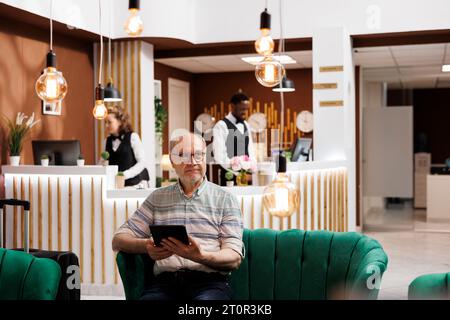 Personne âgée caucasienne assis sur le canapé du hall de l'hôtel, tenant la tablette numérique. Vieil homme attendant dans le salon luxueux et utilise un appareil intelligent pour naviguer sur Internet. Ambiance luxueuse et relaxante. Banque D'Images