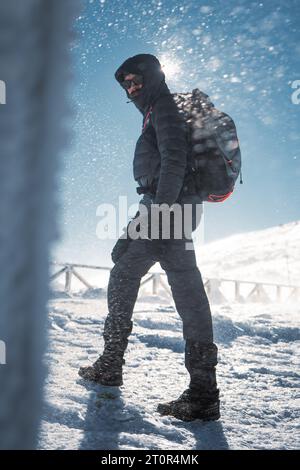 Un randonneur solitaire en vêtements chauds, dans le froid hivernal marche sur la montagne gelée, par jour ensoleillé avec ciel bleu clair et vent fort Banque D'Images