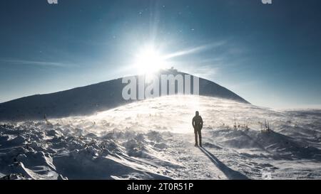 Un randonneur solitaire en vêtements chauds, dans le froid hivernal marche sur la montagne gelée, par jour ensoleillé avec ciel bleu clair et vent fort Banque D'Images