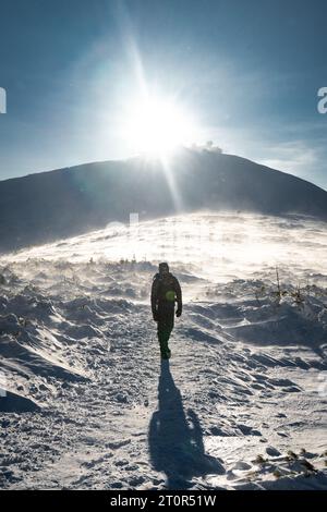 Un randonneur solitaire en vêtements chauds, dans le froid hivernal marche sur la montagne gelée, par jour ensoleillé avec ciel bleu clair et vent fort Banque D'Images