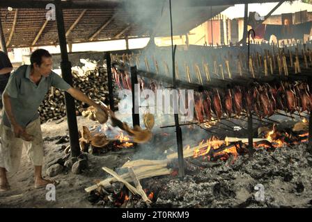 Un travailleur maintient la distribution égale des coques de noix de coco qui sont utilisées comme combustible pour fumer des viandes de thon listao dans une industrie domestique à Bitung, Sulawesi du Nord, en Indonésie. Les contributions des connaissances autochtones à l'innovation technologique offrent un large éventail d'options pour la gestion, entre autres, de la sécurité alimentaire, selon le rapport de 2023 du Groupe d'experts intergouvernemental sur l'évolution du climat (GIEC). Populairement connu sous le nom de cakalang fufu, le thon fumé est considéré comme un aliment indigène exotique et pourrait devenir une solution alternative pour faire face aux impacts du changement climatique sur l’océan. Banque D'Images