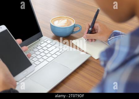 Gros plan de la main masculine écrivant sur le carnet de notes avec stylo, montrant l'écran vierge de l'ordinateur portable et smartphone. Travailler dans le concept de café avec une tasse de café Banque D'Images