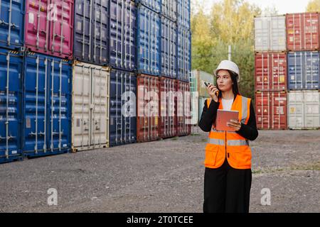 Jeune femme ingénieur portant un casque blanc parlant radio téléphone ou talkie-walkie dans l'entrepôt de conteneurs. Banque D'Images