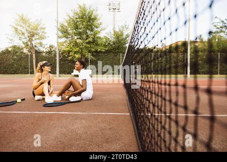 Deux jeunes belles femmes ayant une conversation assise sur un court de tennis à côté du filet. Deux joueurs de tennis amateurs faisant une pause pendant un tennis Banque D'Images