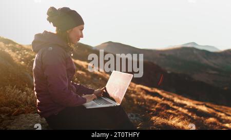 Femme de montagne de coucher de soleil d'automne à distance travaillant sur ordinateur portable. Fille de touriste tapant sur le cahier assis sur la prairie d'herbe orange dans la lumière douce du soleil. Randonnée, trekking, voyage de style de vie en plein air sur les vacances Banque D'Images