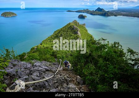 Jeune homme escaladant la montagne rocheuse contre la mer bleue avec la forêt verte Banque D'Images