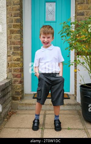 Heureux et excité petit garçon de 4 ans devant la porte d'entrée de sa maison sur son premier jour d'école portant son uniforme et un pantalon court, Royaume-Uni Banque D'Images