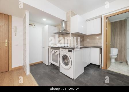 Appartement avec cuisine ouverte équipée sur le coin avec meubles blancs, appareils blancs intégrés, comptoir gris foncé et sol assorti Banque D'Images