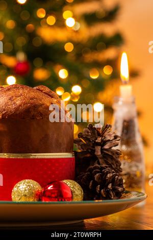 Goiânia, Goias, Brésil – 08 octobre 2023 : un panettone, des pommes de pin, des bougies allumées, des boules et un sapin de Noël en arrière-plan avec des lumières bokeh. Banque D'Images