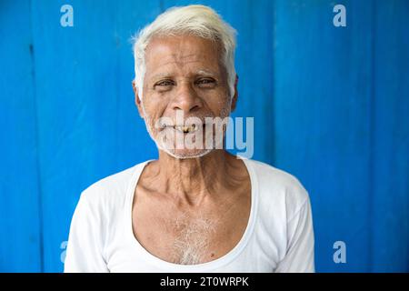 Homme local souriant à la caméra devant un mur en bois bleu dans sa maison dans un petit village sur l'île de Majuli, rivière Brahmapoutre, Assam, Inde Banque D'Images