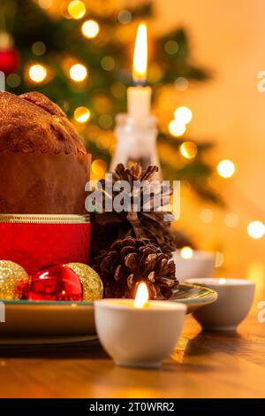 Goiânia, Goias, Brésil – 08 octobre 2023 : un panettone, des pommes de pin, des bougies allumées, des boules et un sapin de Noël en arrière-plan avec des lumières bokeh. Banque D'Images