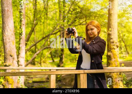Femme photographe avec une veste noire profitant dans un parc d'automne, prenant des photos au coucher du soleil sur un pont en bois Banque D'Images