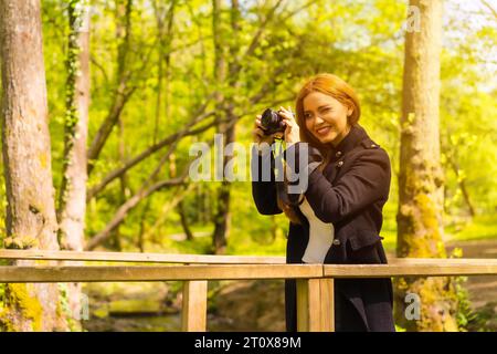 Femme photographe avec une veste noire profitant dans un parc d'automne, prenant des photos au coucher du soleil sur un pont en bois Banque D'Images