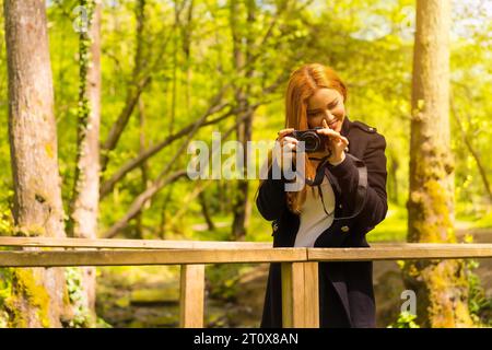 Femme photographe avec une veste noire profitant dans un parc d'automne, souriant et appréciant de prendre des photos au coucher du soleil Banque D'Images