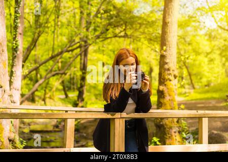 Femme photographe avec une veste noire profitant dans un parc d'automne, prenant des photos sur un pont en bois Banque D'Images