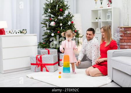 Une famille de trois personnes est assise sur un tapis avec un sapin de Noël en arrière-plan Banque D'Images