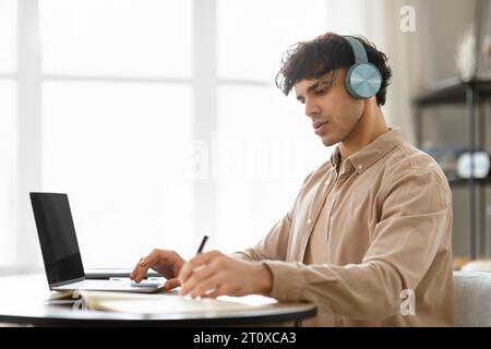 Homme arabe à l'ordinateur portable écrire des notes portant des écouteurs sans fil à l'intérieur Banque D'Images