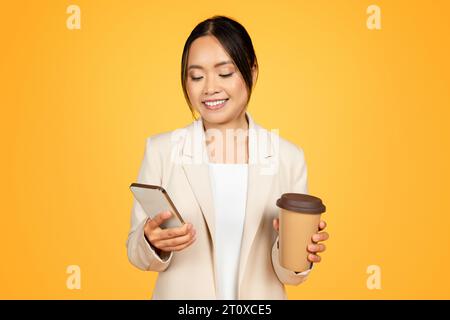 Femme asiatique millénaire concentrée en costume utilisant le téléphone, boire une tasse de café Banque D'Images