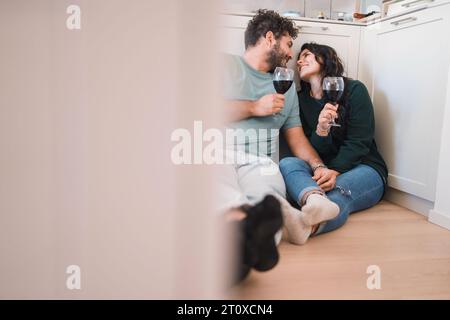 Un jeune couple habillé de façon décontractée, dans leurs années 30, toast avec des verres à vin dans leur nouvelle cuisine. L'homme barbu et la femme aux cheveux foncés, tous deux caucasiens, lovi Banque D'Images