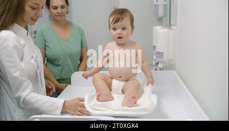 Mère tient bébé sur les mains. Medic met de l'échelle sur une table à langer. Femme médecin pèse enfant dans la chambre pédiatrique moderne. Femme avec fils sur rendez-vous avec pédiatre. Le personnel médical au travail à l'hôpital. Banque D'Images