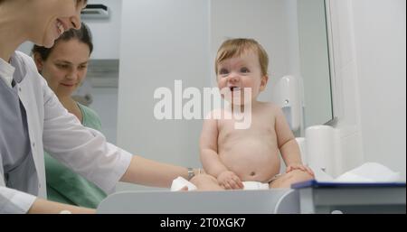 Mère tient bébé sur les mains. Medic met de l'échelle sur une table à langer. Femme médecin pèse enfant dans la chambre pédiatrique moderne. Femme avec fils sur rendez-vous avec pédiatre. Le personnel médical au travail à l'hôpital. Banque D'Images