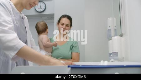 Mère tient bébé sur les mains. Medic met de l'échelle sur une table à langer. Femme médecin pèse enfant dans la chambre pédiatrique moderne. Femme avec fils sur rendez-vous avec pédiatre. Le personnel médical au travail à l'hôpital. Banque D'Images