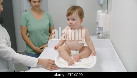 Mère tient bébé sur les mains. Medic met de l'échelle sur une table à langer. Femme médecin pèse enfant dans la chambre pédiatrique moderne. Femme avec fils sur rendez-vous avec pédiatre. Le personnel médical au travail à l'hôpital. Banque D'Images