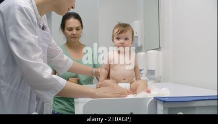 Mère tient bébé sur les mains. Medic met de l'échelle sur une table à langer. Femme médecin pèse enfant dans la chambre pédiatrique moderne. Femme avec fils sur rendez-vous avec pédiatre. Le personnel médical au travail à l'hôpital. Banque D'Images