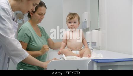 Mère tient bébé sur les mains. Medic met de l'échelle sur une table à langer. Femme médecin pèse enfant dans la chambre pédiatrique moderne. Femme avec fils sur rendez-vous avec pédiatre. Le personnel médical au travail à l'hôpital. Banque D'Images