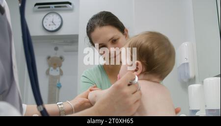 Petit garçon assis sur une table à langer sur rendez-vous avec le pédiatre dans le service d'hôpital léger. Mère tient son bébé. Le médecin féminin utilise le stéthoscope et vérifie l'état de l'enfant. Medic au travail en clinique. Banque D'Images