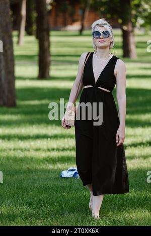 Jeune femme blonde aux cheveux courts se tient à pleine hauteur dans un parc portant des lunettes de soleil et une robe noire. Banque D'Images