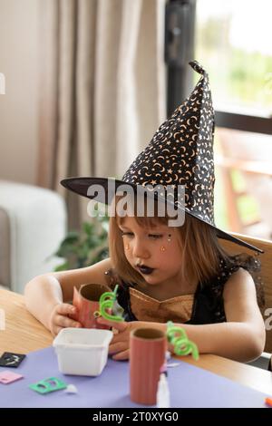 Une fille mignonne dans un costume de sorcière fait un artisanat de papier pour Halloween. Artisanat pour enfants pour Halloween. Banque D'Images