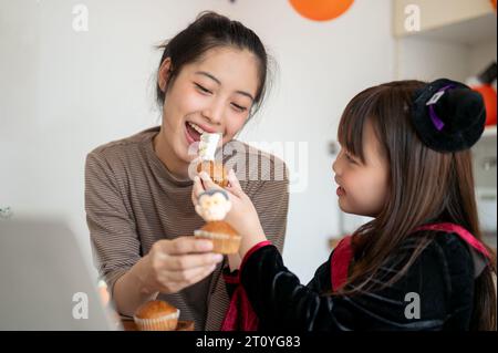 Une jeune maman asiatique attentionnée et belle aime manger des cupcakes Halloween et célébrer Halloween avec sa petite fille à la maison. Banque D'Images