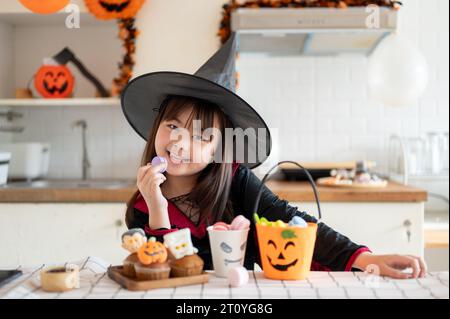 Une jeune fille asiatique mignonne et joyeuse dans un costume d'Halloween s'assoit à une table de cuisine avec des desserts d'Halloween et aime célébrer l'effrayant Hallowee Banque D'Images