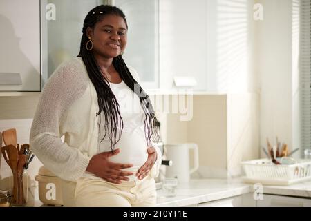 Portrait de femme enceinte souriante positive avec des cheveux tressés debout dans la cuisine Banque D'Images