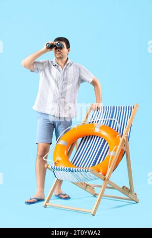 Jeune homme avec des jumelles, bouée annulaire et chaise longue sur fond bleu Banque D'Images