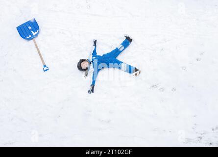 garçon en costume d'hiver bleu déneigeait après le blizzard et s'allongeait sur la neige pour se reposer, avec une grande pelle à proximité. Hiver, mauvais temps, froid, jeu Banque D'Images