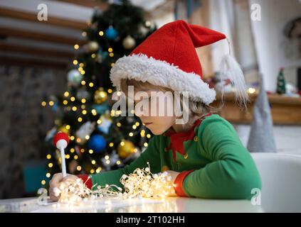 L'enfant s'assoit à une table sur fond de guirlandes de Noël et écrit une lettre au Père Noël. Un garçon de 7 ans portant un chapeau rouge rêve de cadeaux. Cadeau WIS Banque D'Images