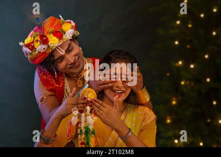 Jeune homme déguisé en Seigneur Krishna et couvrant les yeux de la femme à l’occasion de Janmashtami Banque D'Images