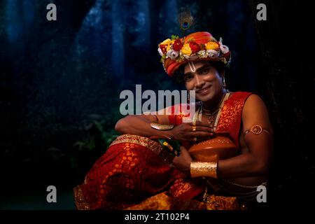 Jeune homme déguisé en Seigneur Krishna et assis avec un pot de beurre à l’occasion de Janmashtami Banque D'Images