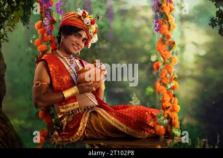 Jeune homme déguisé en Seigneur Krishna et assis sur une balançoire avec un pot de beurre à l’occasion de Janmashtami Banque D'Images