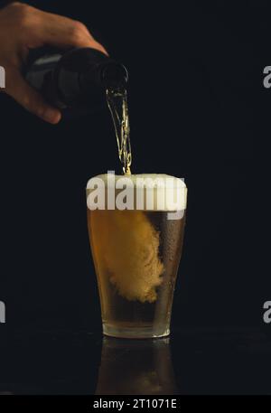 Plan frontal d'un verre de bière et d'une main versant de la bière sur un fond noir avec un jet formant de la mousse. Banque D'Images