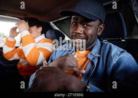 Jeune rappeur afro-américain souriant en casquette et veste en denim regardant la caméra tout en étant assis sur la banquette arrière de la voiture contre son copain Banque D'Images