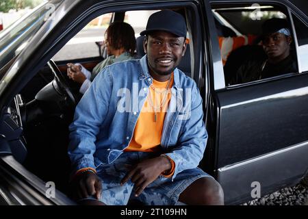 Jeune danseur de hip hop souriant en casquette noire, veste en denim et short assis sur le siège avant de la voiture et regardant la caméra contre ses copains Banque D'Images